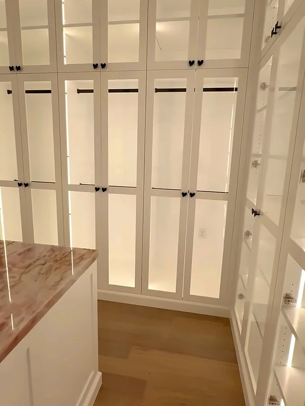 Wall of white glass-front closet cabinets with interior LED lighting and hanging rods, marble island corner in front