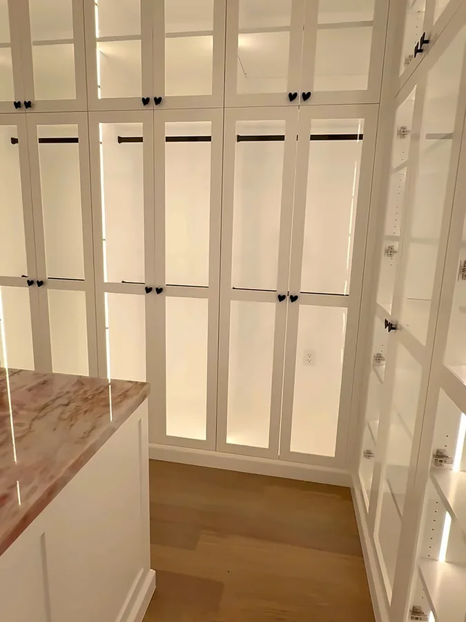 Wall of white glass-front closet cabinets with interior LED lighting and hanging rods, marble island corner in front