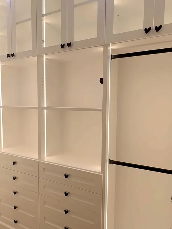 White closet wall with glass-front upper cabinets over lit open shelves, a drawer bank and long hanging