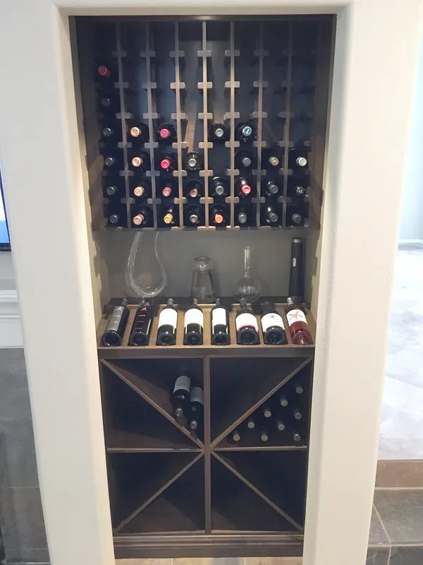 Dark-stained built-in wine tower in a wall niche, with bottle cubbies, a display shelf and an X-shaped bin below, Newbury Park